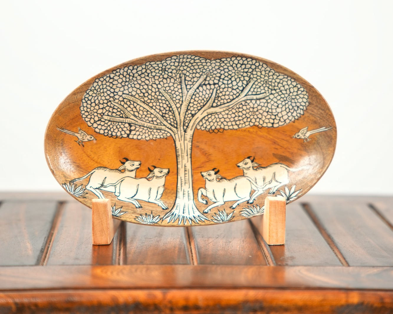 An oval wooden decorative plate with hand-carved animal and tree motifs, displayed on a small wooden stand on a wooden table against a white background.