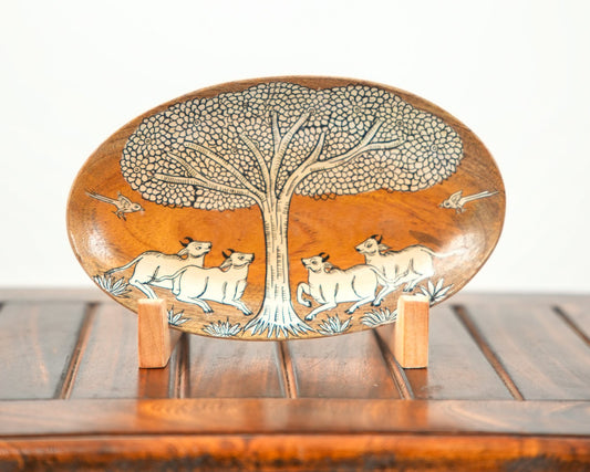 An oval wooden decorative plate with hand-carved animal and tree motifs, displayed on a small wooden stand on a wooden table against a white background.
