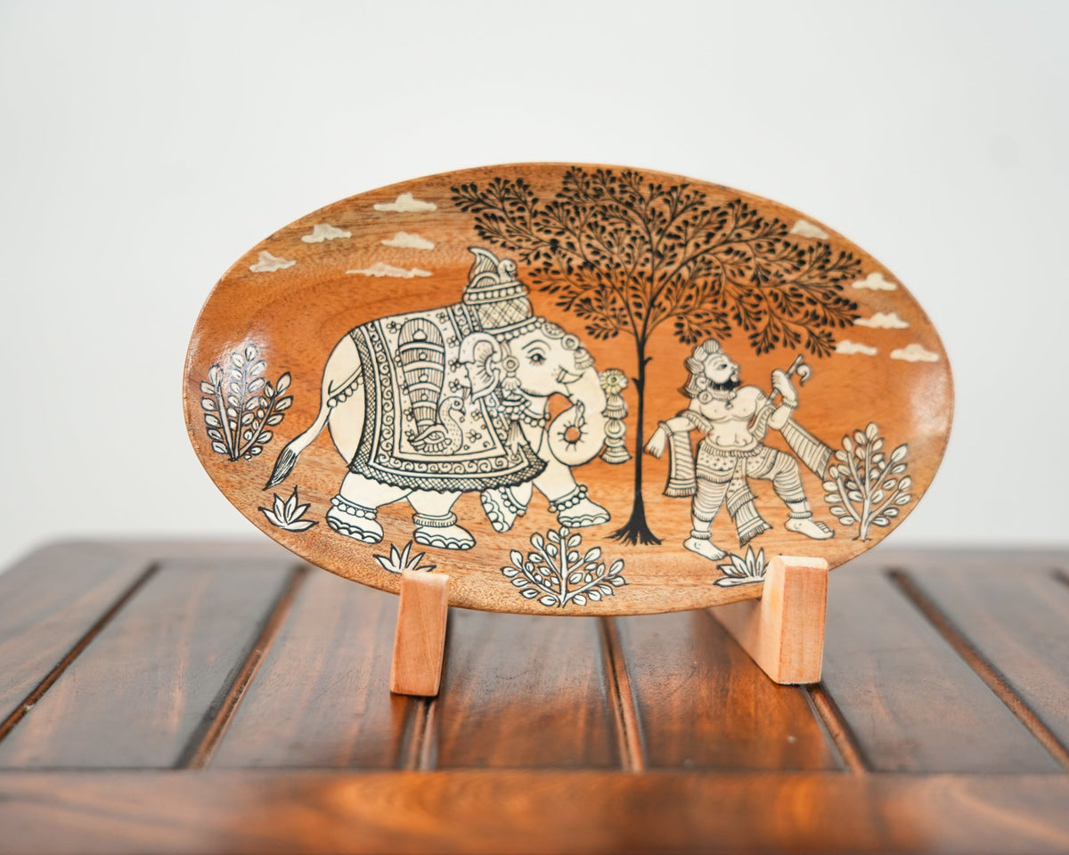 An oval hand-painted clay plate featuring an elephant and a man under a tree, displayed on a small wooden stand on a dark wooden table against a light gray background.