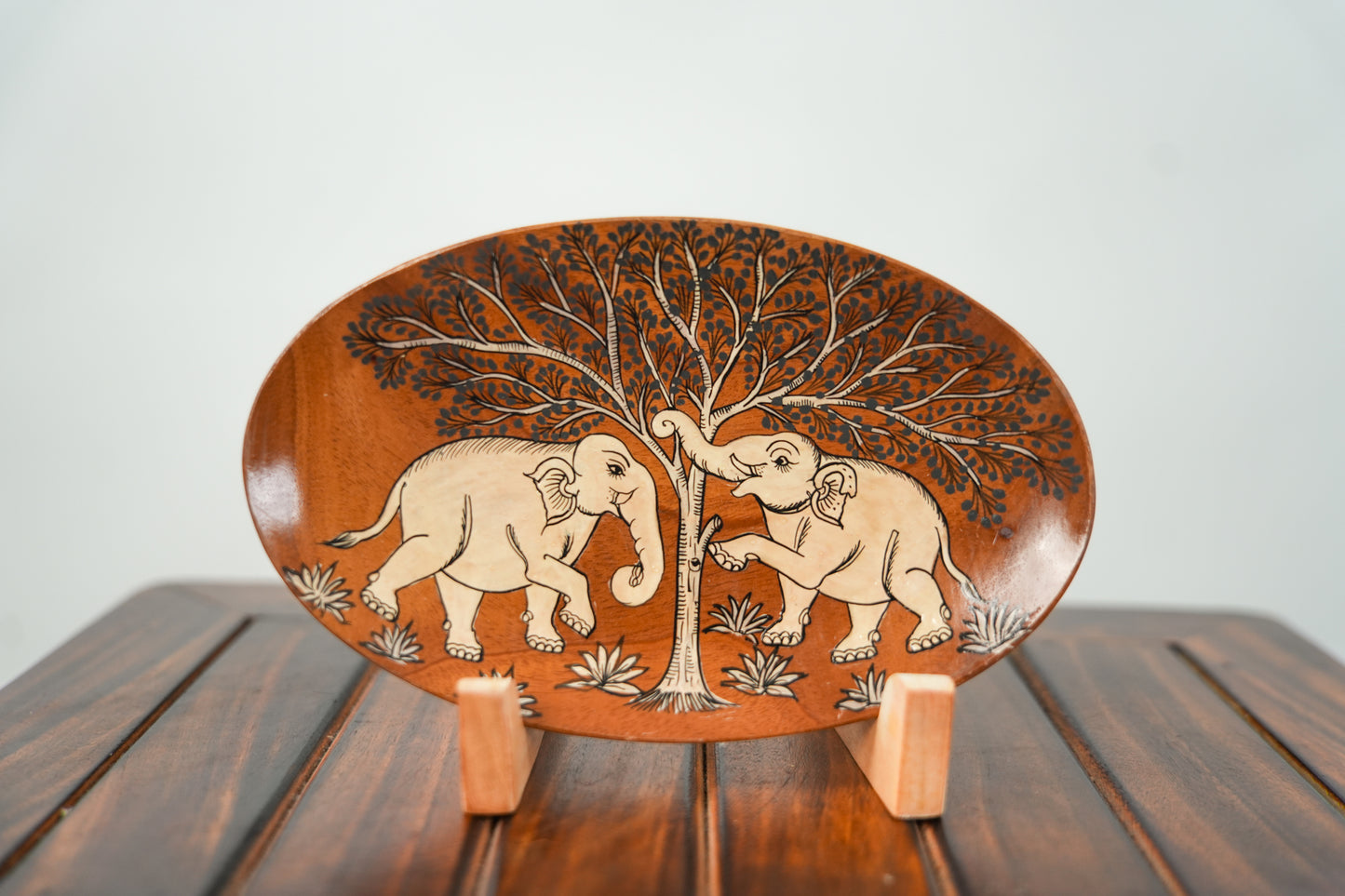 An oval wooden decorative plate with hand-carved elephant and tree designs, displayed on a small wooden stand over a dark wooden table against a light gray background.