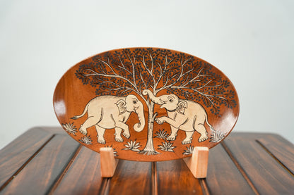 An oval wooden decorative plate with hand-carved elephant and tree designs, displayed on a small wooden stand over a dark wooden table against a light gray background.