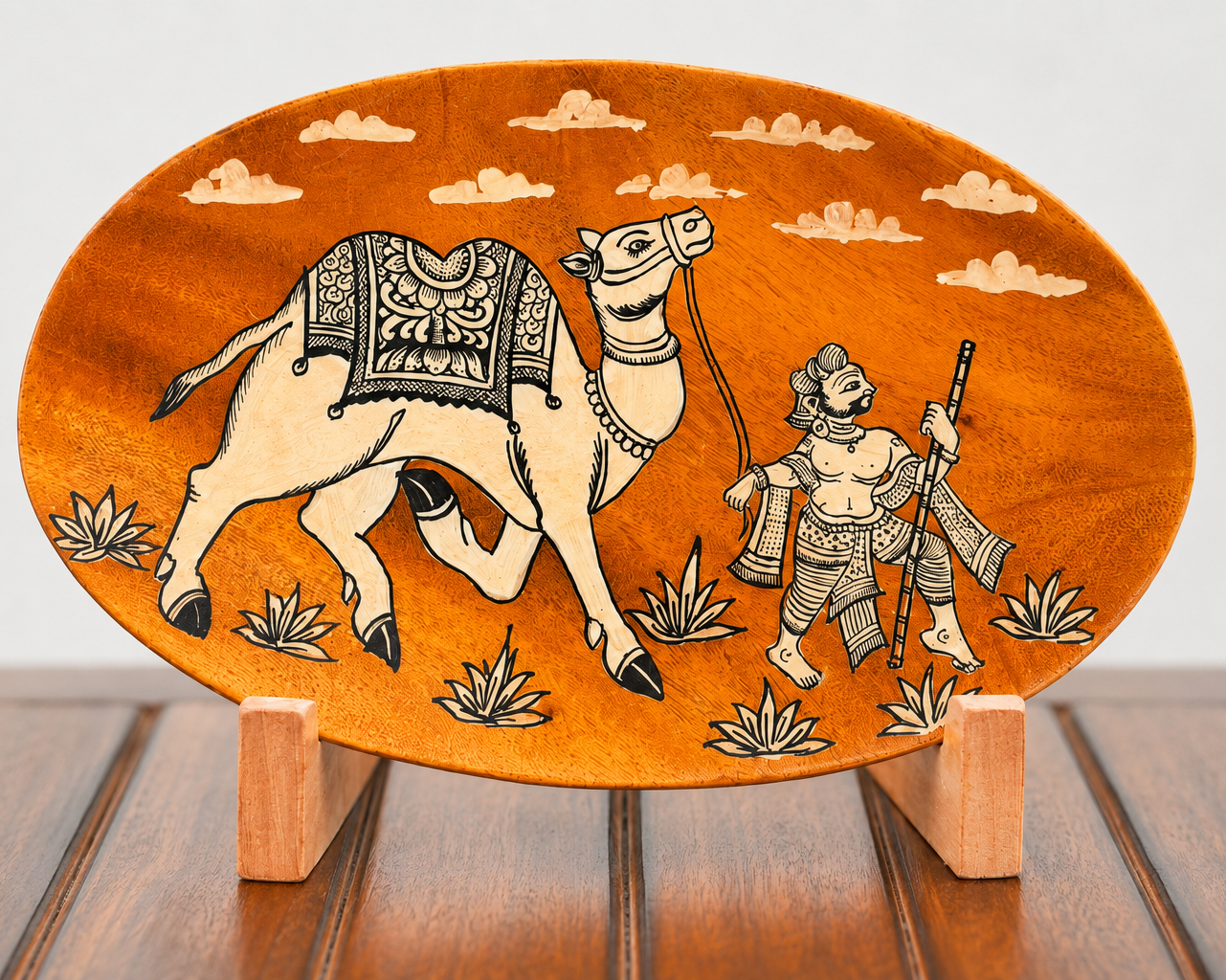 An oval wooden decorative plaque featuring an engraved camel and warrior scene, displayed on small wooden stands over a dark wood surface against a light gray background.